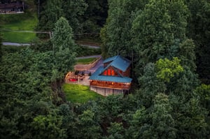 Aerial view showcases a charming log cabin with a distinctive blue metal roof and red accents, nestled privately among lush forest greenery with an inviting deck for outdoor relaxation.