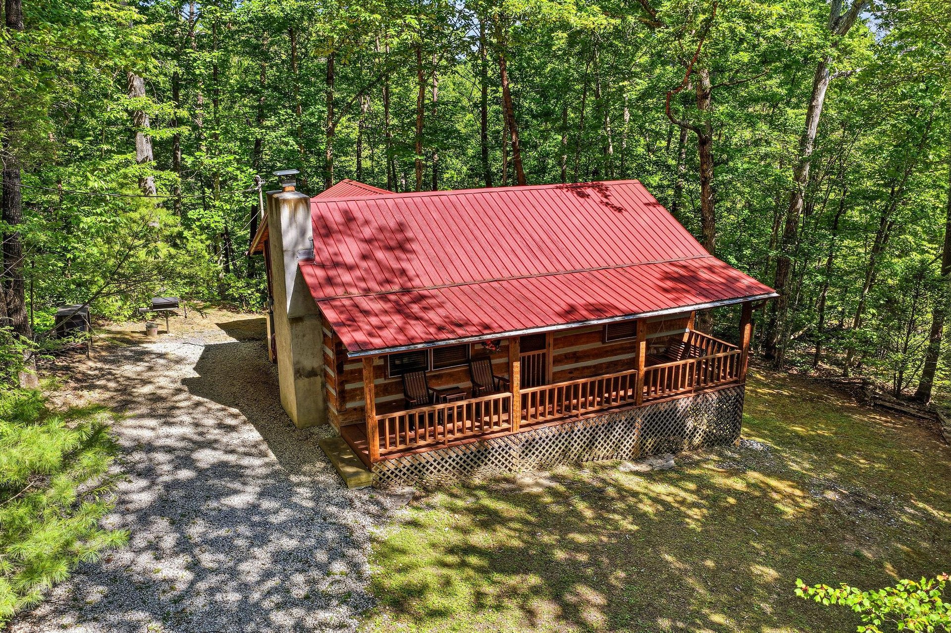 A charming log cabin with a distinctive red roof nestled among lush forest trees, offering a peaceful wooded retreat with convenient parking and outdoor grilling space.