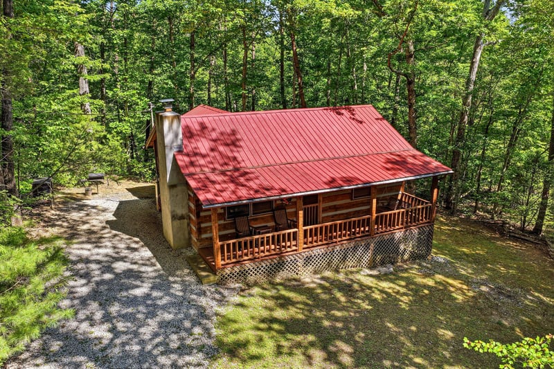 A charming log cabin with a distinctive red roof nestled among lush forest trees, offering a peaceful wooded retreat with convenient parking and outdoor grilling space.