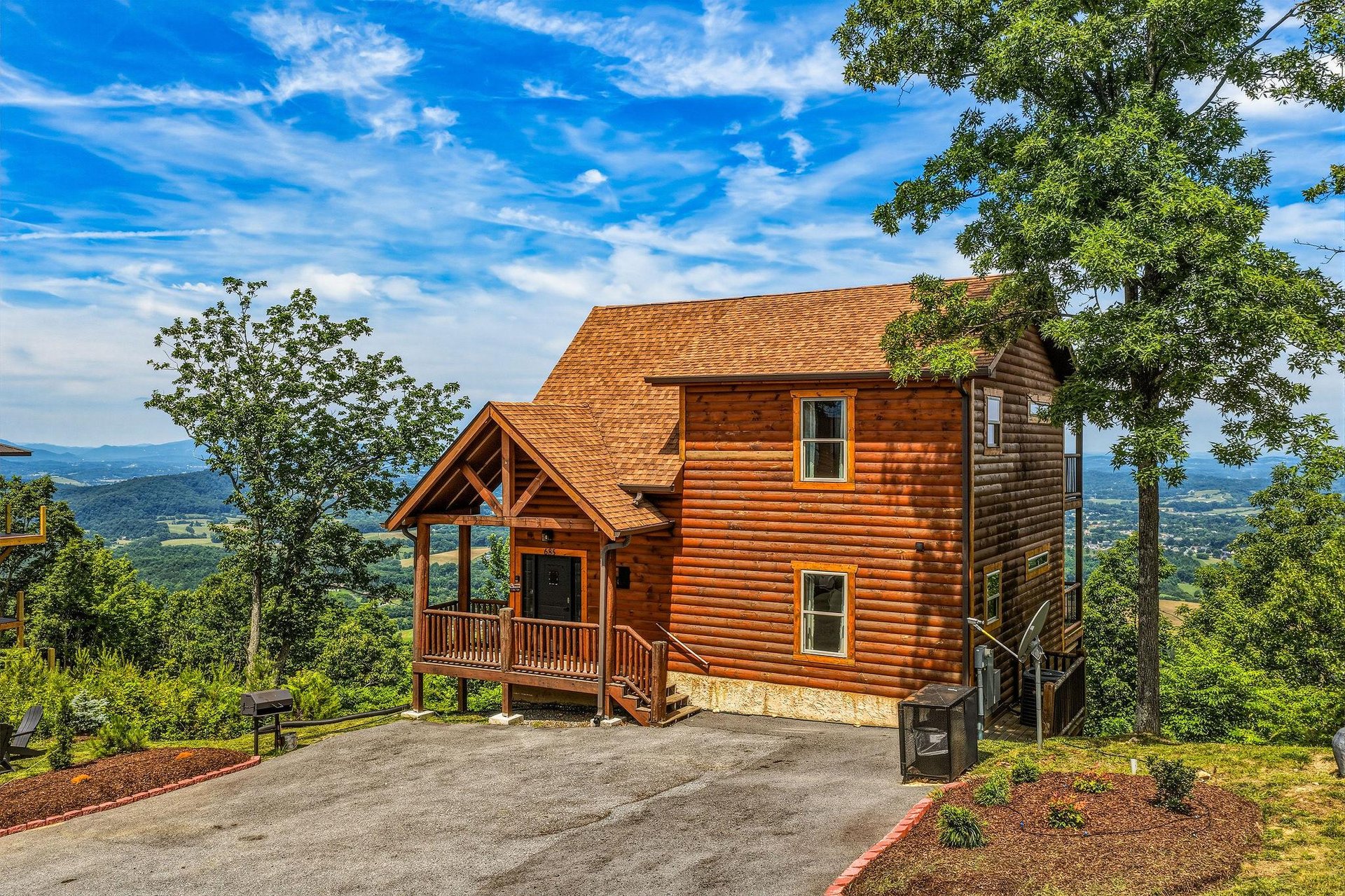 A charming log cabin perched on a mountainside, featuring warm wood siding, a welcoming covered entry deck, and sweeping valley views. Surrounded by natural landscape and mature trees.