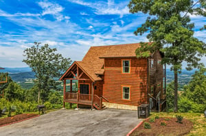 A charming log cabin perched on a mountainside, featuring warm wood siding, a welcoming covered entry deck, and sweeping valley views. Surrounded by natural landscape and mature trees.