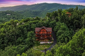 Aerial view of a secluded log cabin nestled in lush forested mountains at dusk, showcasing the propertys private natural setting and scenic landscape.