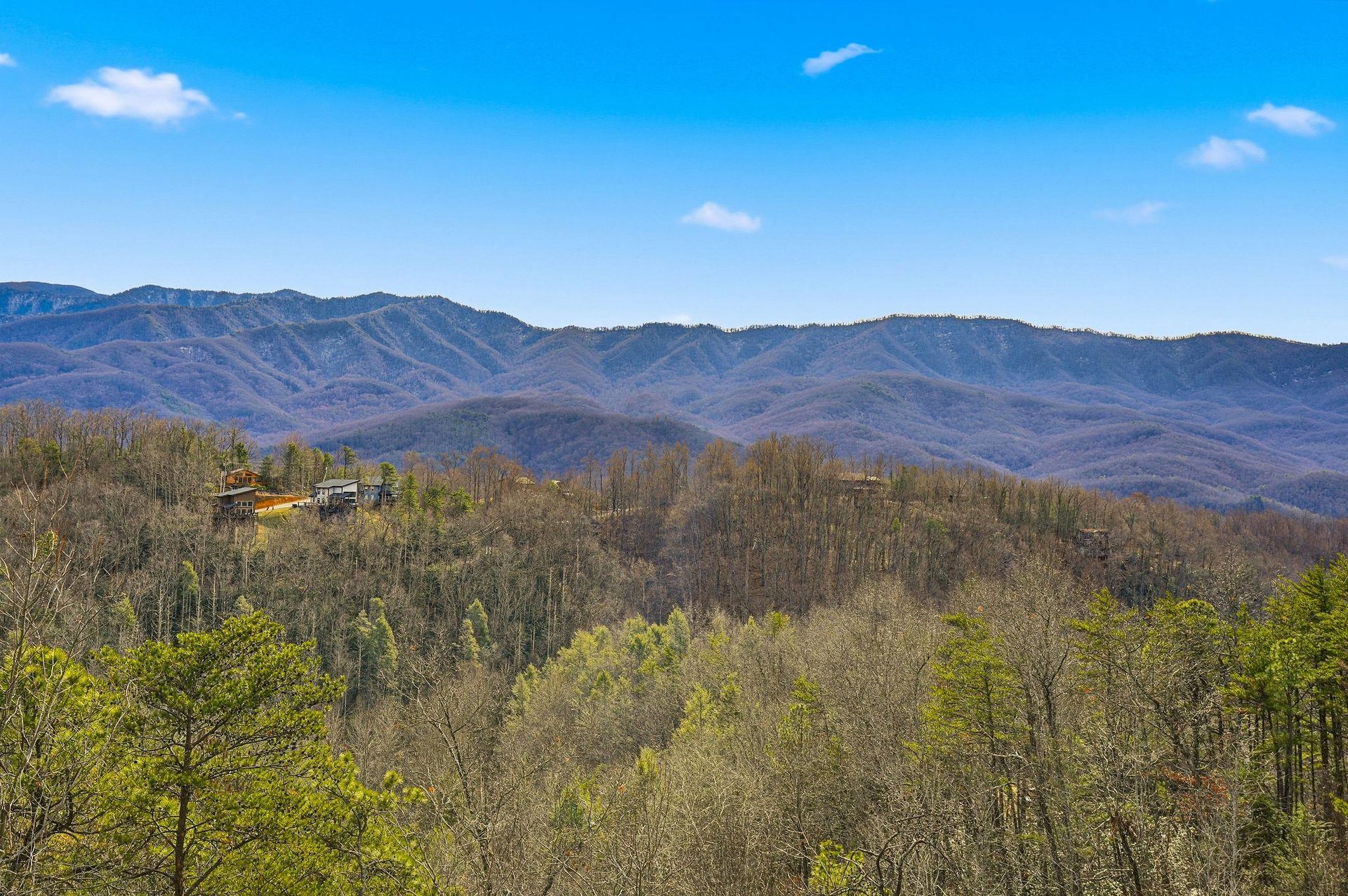Stunning mountain views stretch across layered ridges beneath bright blue skies, with a charming cabin nestled among the forested hillsides.