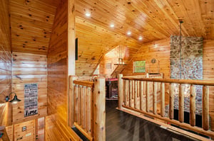 Cozy log railings and warm wood paneling surround your loft space, with rustic stone fireplace adding cabin charm. Natural light filters through, inviting you to unwind in this inviting retreat.