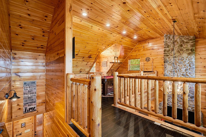 Cozy log railings and warm wood paneling surround your loft space, with rustic stone fireplace adding cabin charm. Natural light filters through, inviting you to unwind in this inviting retreat.