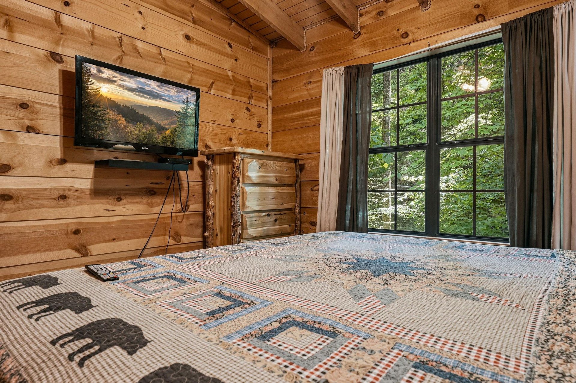 Wake in your cozy cabin bedroom with forest views through expansive windows, where rustic woodbeamed ceilings and natural pine walls create your perfect mountain retreat.