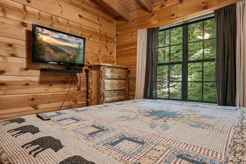Wake in your cozy cabin bedroom with forest views through expansive windows, where rustic woodbeamed ceilings and natural pine walls create your perfect mountain retreat.