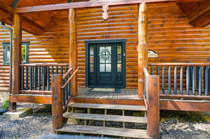 Charming log cabin entrance with welcoming green door and wraparound porch invites you to your mountain retreat.