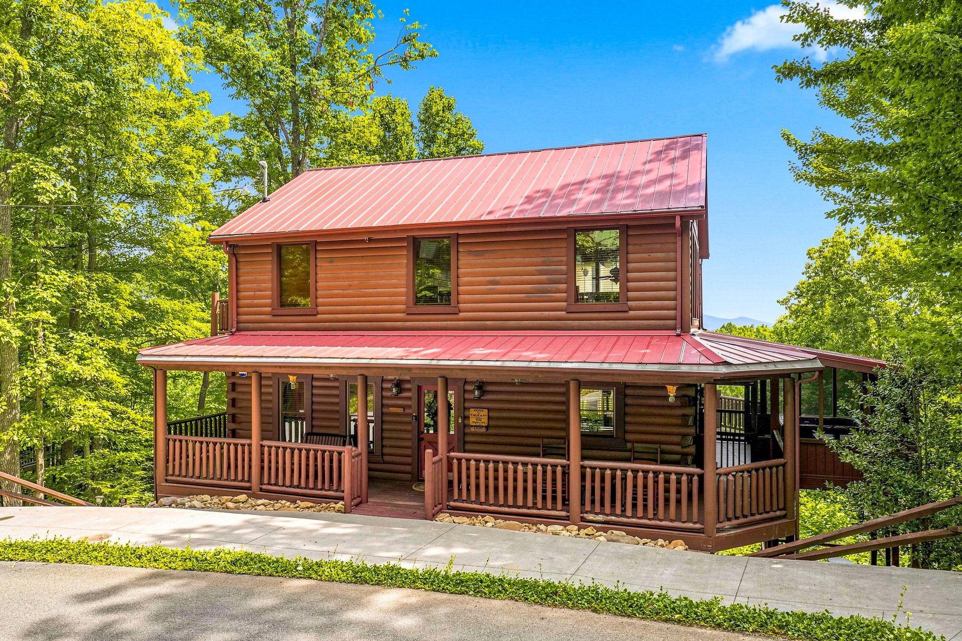 A charming twostory log cabin with a wraparound porch nestled among towering trees, offering a peaceful mountain retreat with ample parking and natural surroundings.