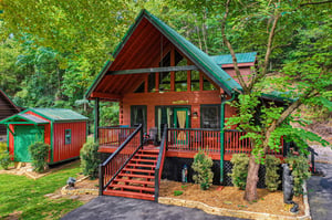 Charming Aframe mountain cabin with welcoming front porch, stained wood siding, green metal roof, nestled among mature trees with beautifully landscaped grounds.