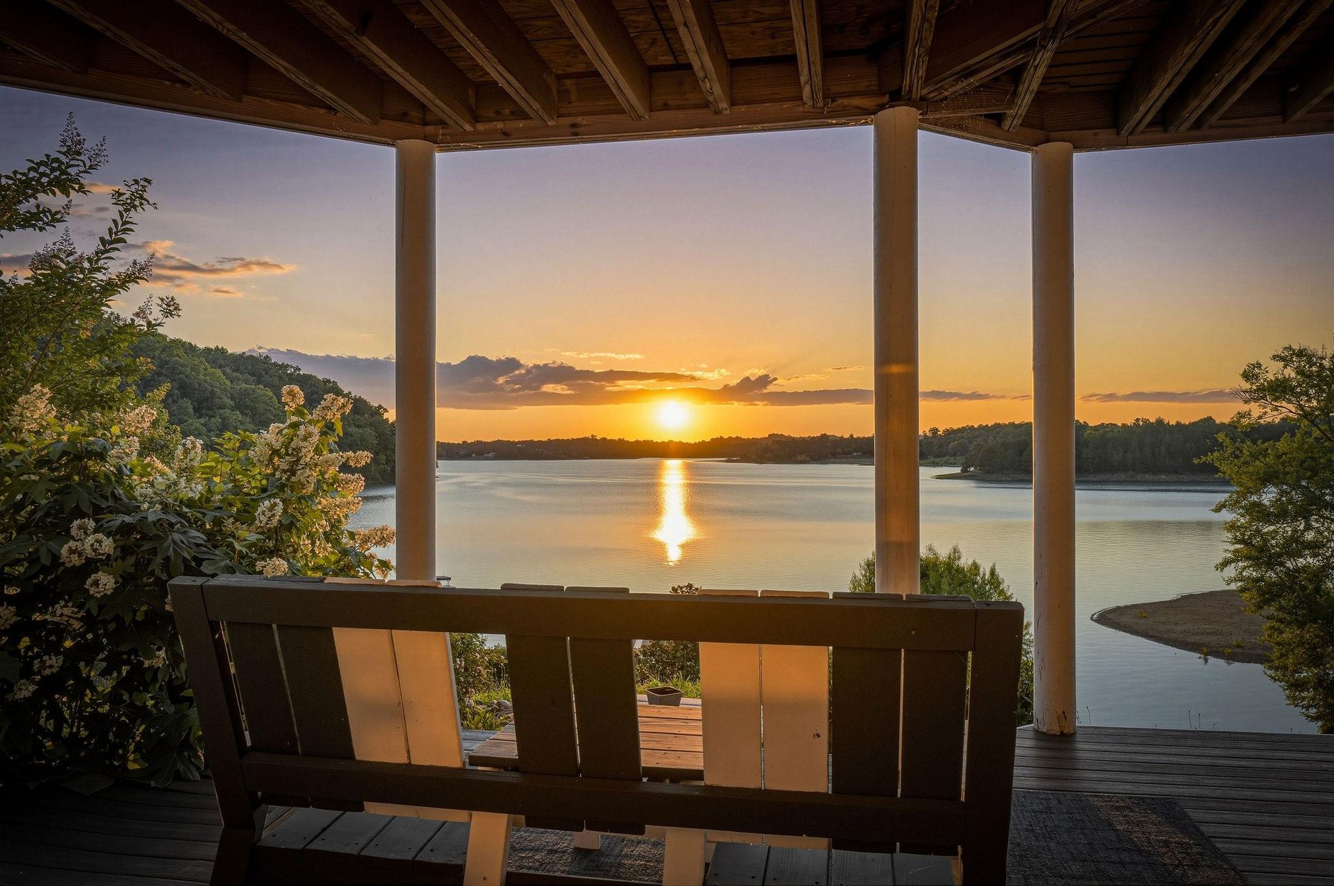 A covered deck frames a tranquil lake as the sun sets over the water and surrounding hills, creating a peaceful arrival scene for the property.