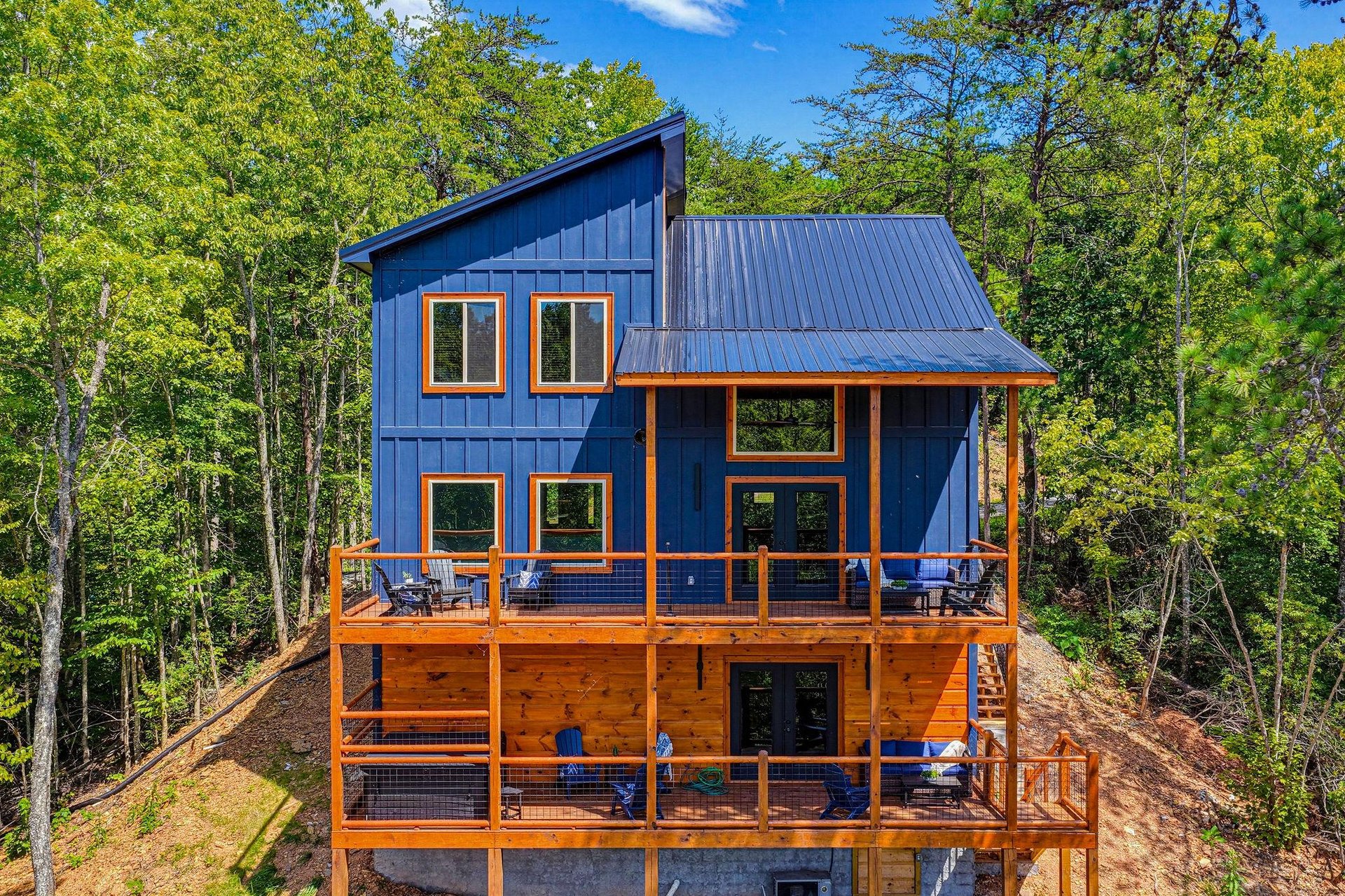 This striking contemporary cabin features bold blue siding with warm cedar accents and expansive multilevel decks tucked into a lush forest setting under clear blue skies.