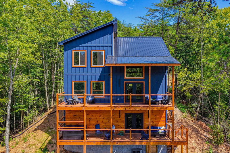 This striking contemporary cabin features bold blue siding with warm cedar accents and expansive multilevel decks tucked into a lush forest setting under clear blue skies.
