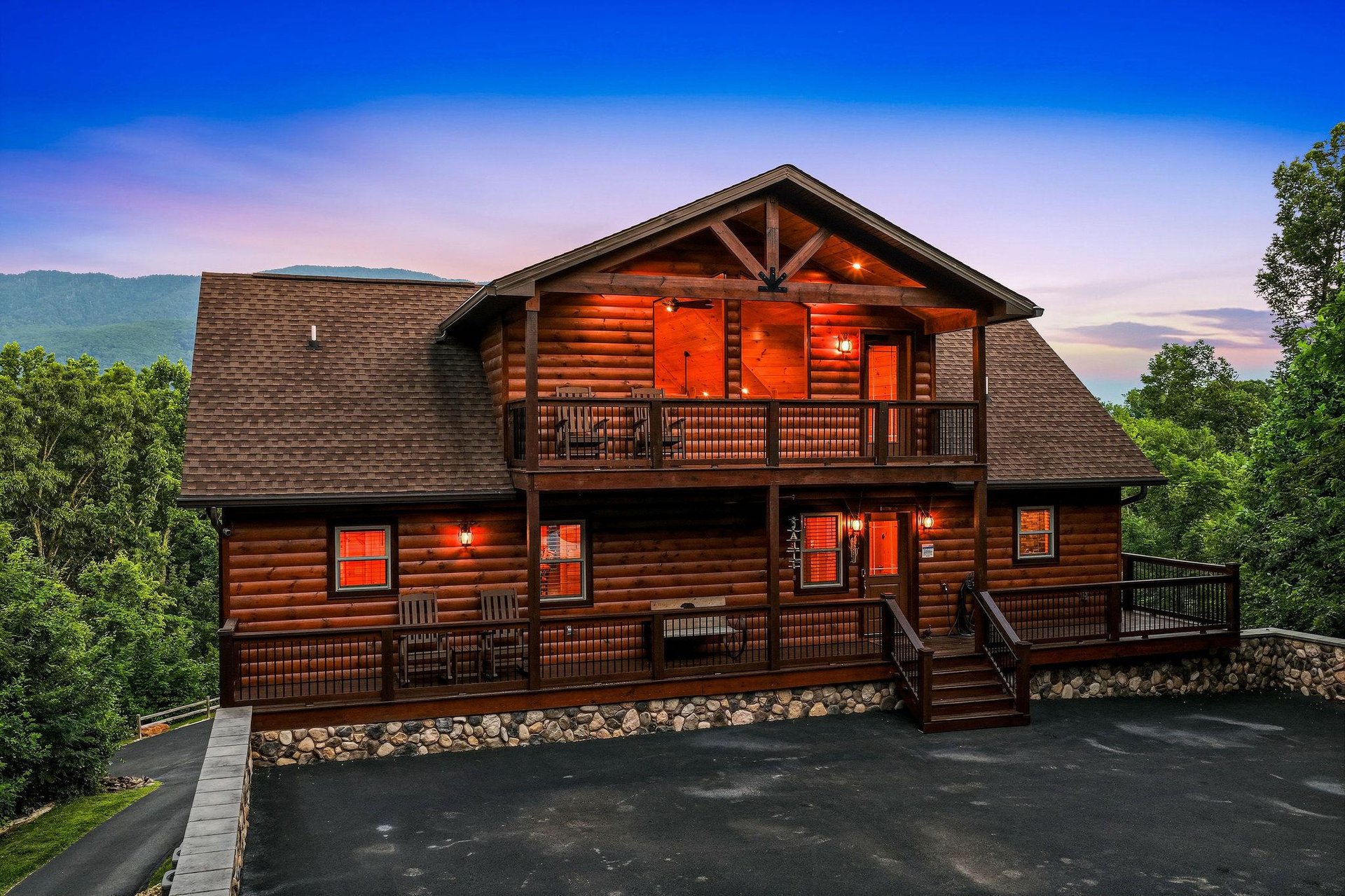 A stunning log cabin rental framed by lush forest and distant mountains, glowing warmly at dusk with spacious decks inviting arrival.