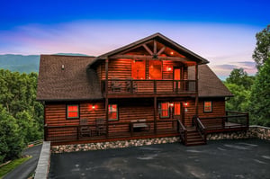 A stunning log cabin rental framed by lush forest and distant mountains, glowing warmly at dusk with spacious decks inviting arrival.