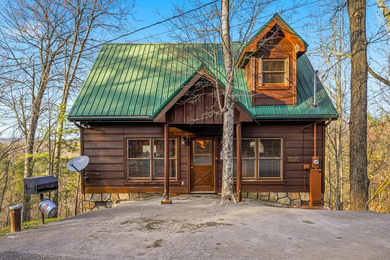Charming log cabin with green metal roof nestled among trees, featuring covered entrance and private parking area.