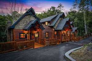 This stunning mountain lodge features warm wood exteriors and multiple private decks, nestled among towering trees with a breathtaking sunset sky as your backdrop.