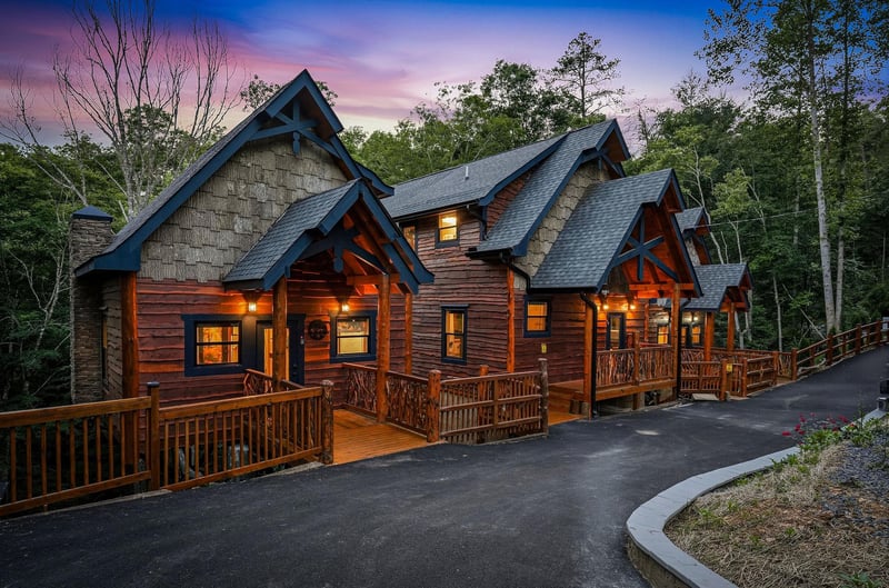 This stunning mountain lodge features warm wood exteriors and multiple private decks, nestled among towering trees with a breathtaking sunset sky as your backdrop.