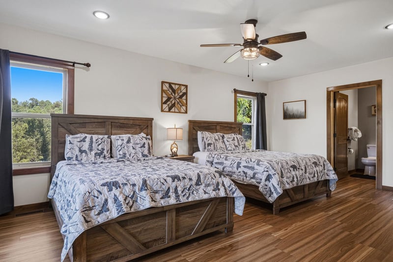 Unwind in your spacious bedroom with two cozy beds, warm wood finishes, and a ceiling fan for comfort. Natural light pours through large windows showcasing treetop views.