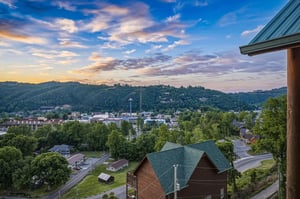 Elevated vantage point overlooking the Gatlinburg area at dusk, showcasing surrounding mountain ridges, forested hillsides, and the towns layout below. The propertys deck provides this panoramic per