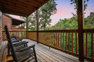 A peaceful covered deck framed by forest greenery offers a serene spot to unwind with mountain breezes and soft pastel skies at dusk.
