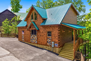 Charming log cabin nestled among towering trees with distinctive green metal roofing and inviting front porch steps.