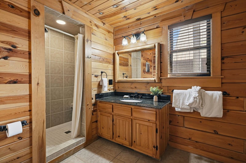 Unwind in your rustic cabin bathroom, where warm wood surrounds granite counters and a spacious tiled shower awaits your refreshing retreat.