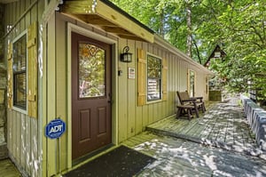 Sunlight filters through towering trees onto this charming cabin entrance, where a welcoming porch and peaceful wooded surroundings invite your arrival.