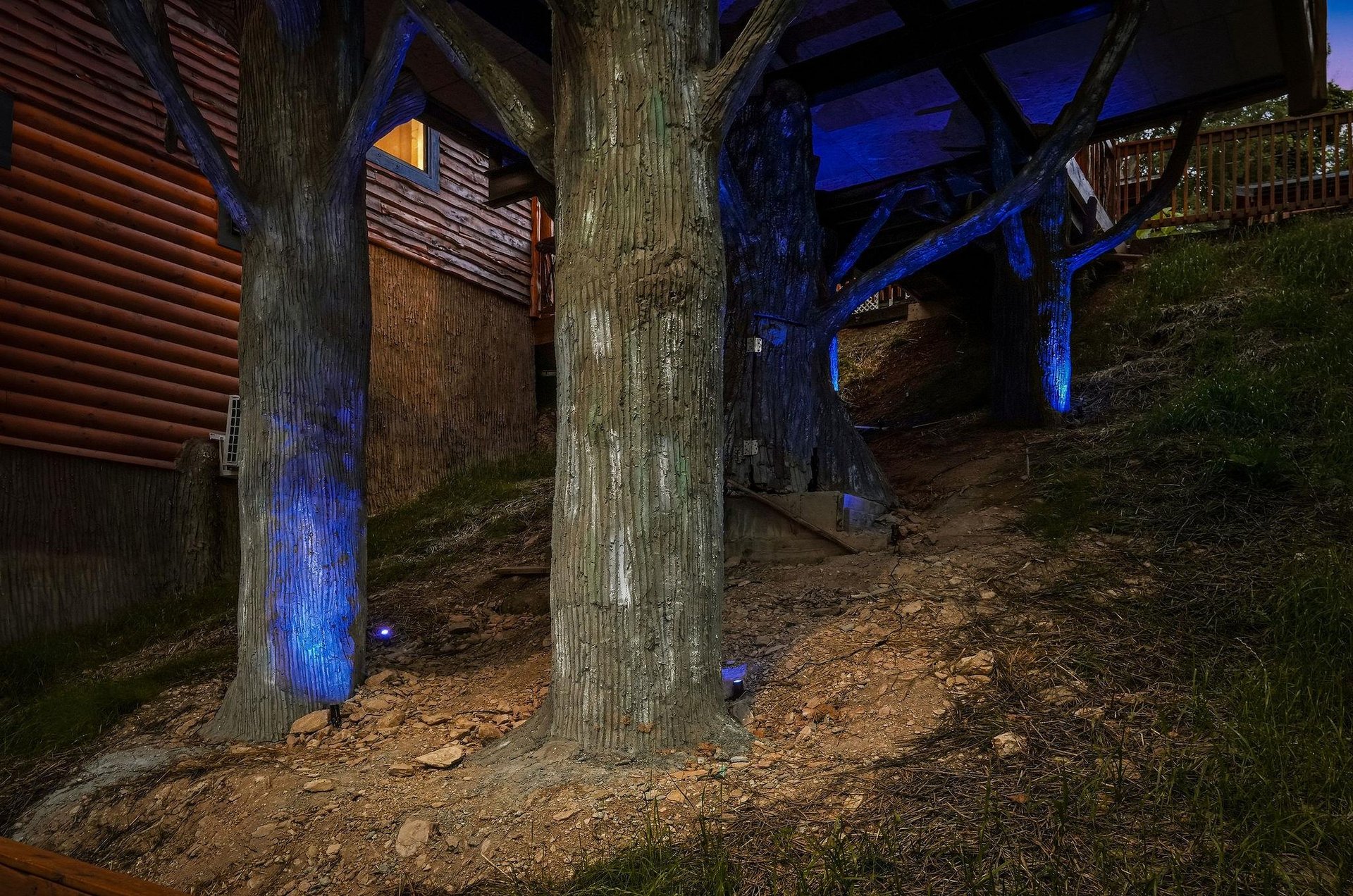 A woodsy cabin retreat illuminated by moonlight and decorative blue accent lighting, nestled among mature trees on a sloping hillside.