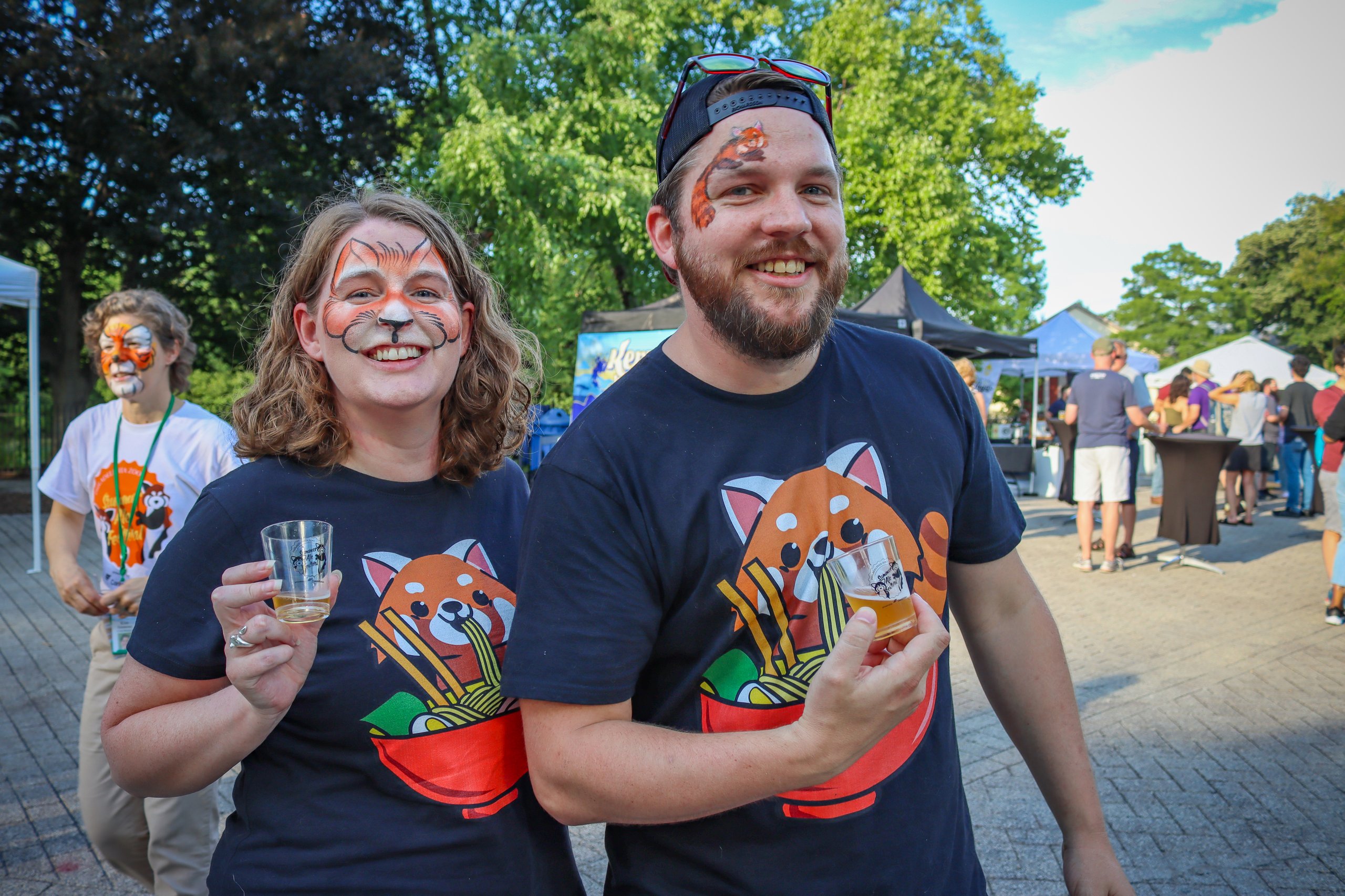 A couple poses for a photo holding branded Summer Ale Festival shot glasses. Their faces are painted to look like tigers.