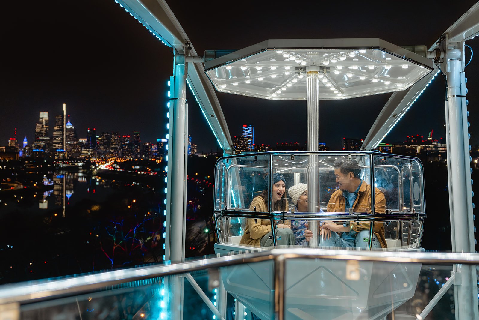 A family of three rides in a ferris wheel gondola, the Philadelphia city skyline lit up in the evening sky behind them.
