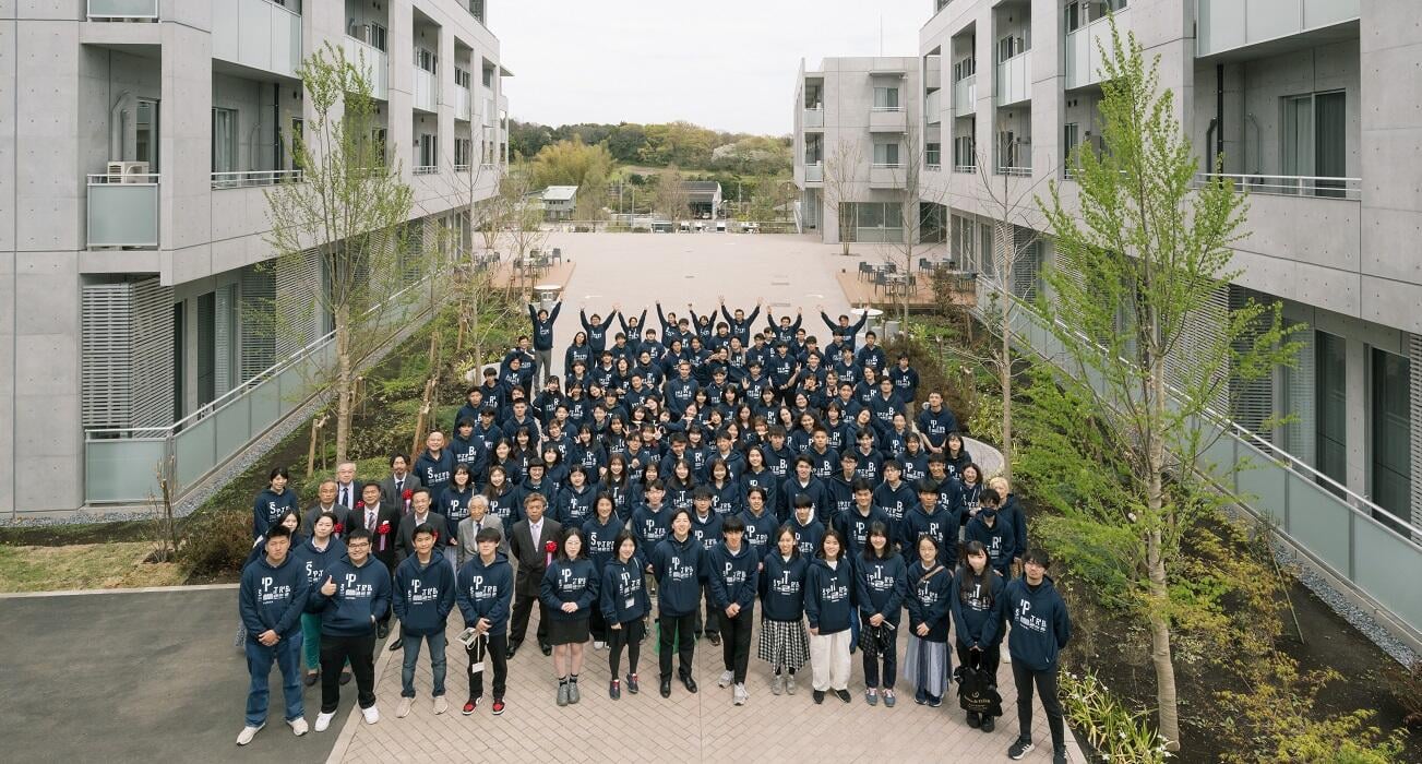 コンクリート造のモダンな建物の間にある広場で、お揃いのネイビーのパーカーを着た大勢の若者たちが集合写真を撮っている様子。前列にはスーツ姿の男性数名も並んでおり、後列の人々は両手を上げてポーズをとっている。