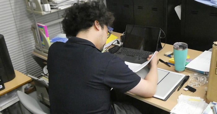 Teachers judging thesis-style works (large quantities of works are visible on desks and shelves)