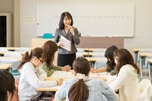 ホワイトボードの前に立ち、机を囲んで座る学生たちに笑顔で話しかける女性講師。