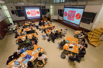 Students working in small groups at tables during a class session in a large classroom.