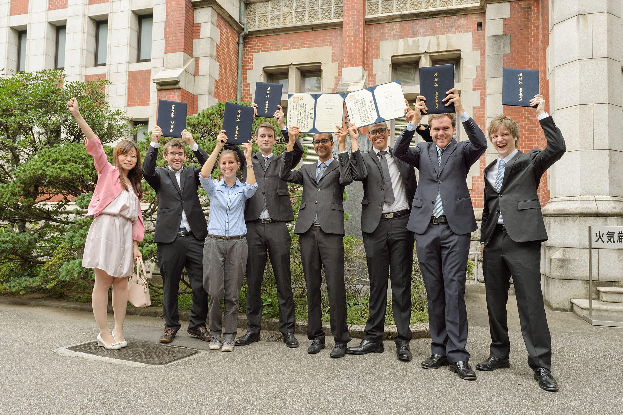 Eight graduates in professional attire celebrate in front of a red brick building, smiling and holding up blue diploma folders in achievement.