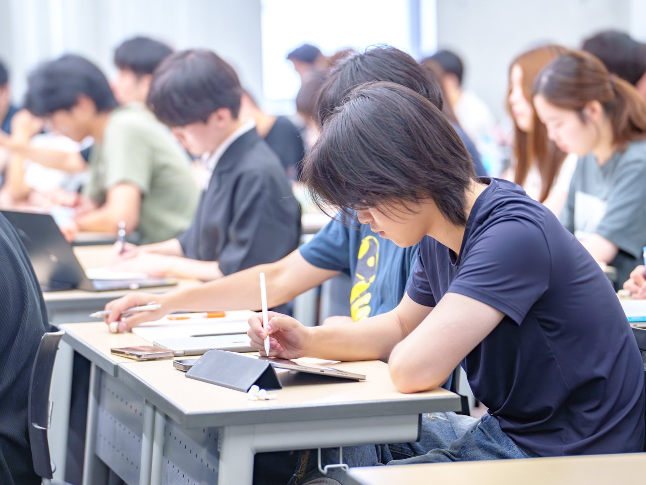 Students working diligently on their assignments. On their desks, they can be seen studying in their own individual styles, such as using PCs or writing on paper.