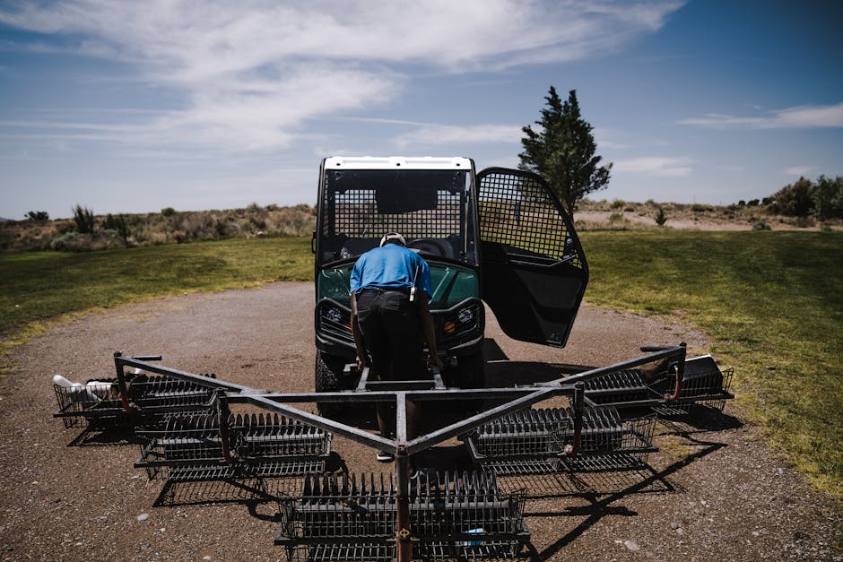 A worker maintaining golf course equipment with a tractor under clear skies.