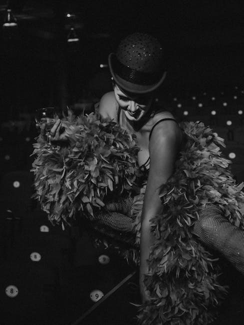Black and white photo of a performer in feathers, holding a drink, exuding glamour on stage.
