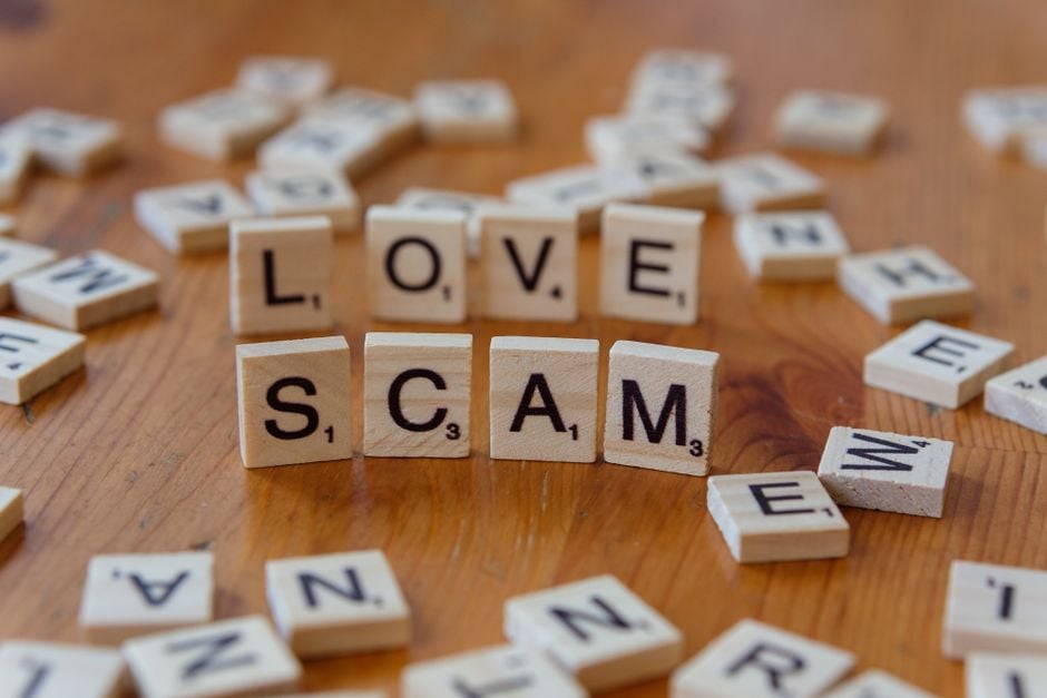 Scrabble tiles form words 'LOVE' and 'SCAM' on a wooden surface, illustrating deceit in relationships.