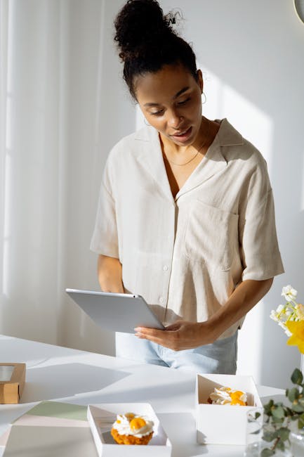 Black businesswoman in casual attire looks at tablet while planning cupcake designs in bright indoor setting.