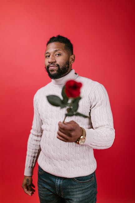 Smiling man in sweater holds a red rose on a vibrant background, perfect for Valentine's Day.