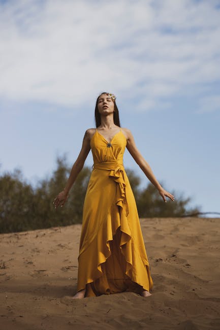 A serene woman meditating in a yellow dress on a sandy hill under a blue sky.