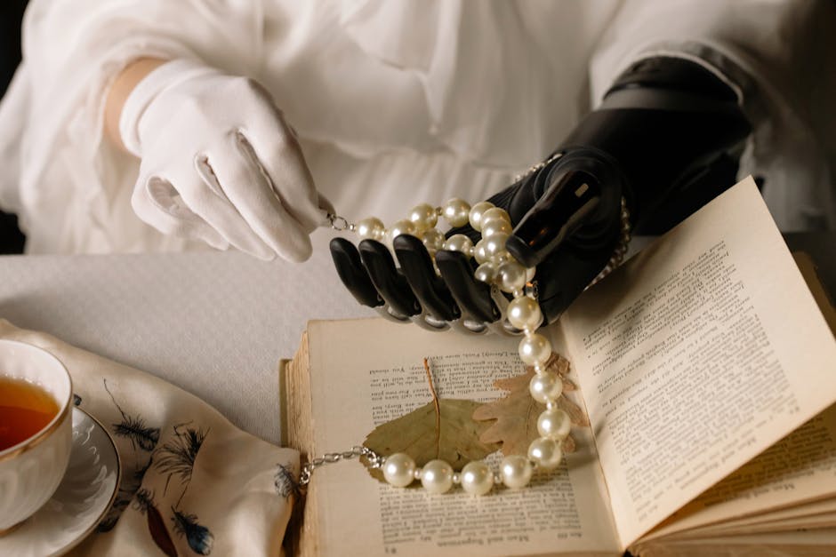 A graceful prosthetic hand holds a pearl necklace over an open book, blending technology and elegance.