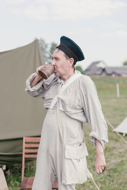 A man in historical clothing drinks from a mug near a tent in a field.