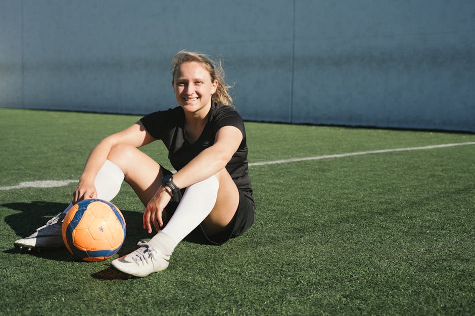 A smiling female soccer player sitting on a soccer field with a ball, enjoying a sunny day.