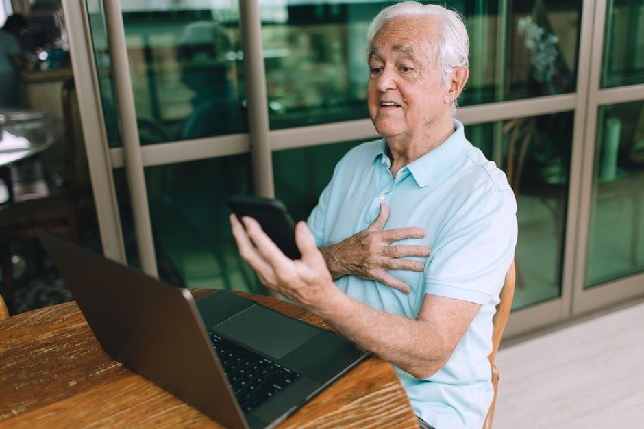 Elderly man interacting with technology using a smartphone and laptop indoors.