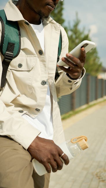 Young man with backpack using cellphone while walking outdoors on a sunny day.