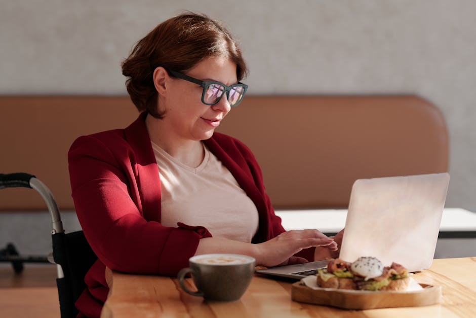 Woman in a red blazer working on laptop at café table with coffee and snack.
