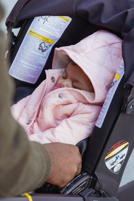 A baby peacefully sleeping in a stroller, covered in a pink jacket. Outdoor scene.